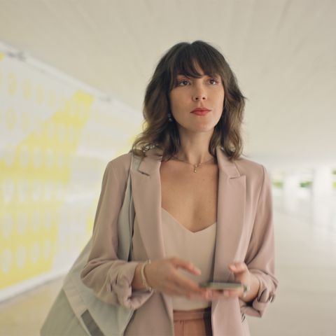 Uma mulher elegante caminha por um corredor olhando em frente com determinação. A parede da esquerda possui curvas amarelas. A mulher veste blêizer na cor bege, segura uma bolsa embaixo do braço esquerdo e segura um celular com as duas mãos.