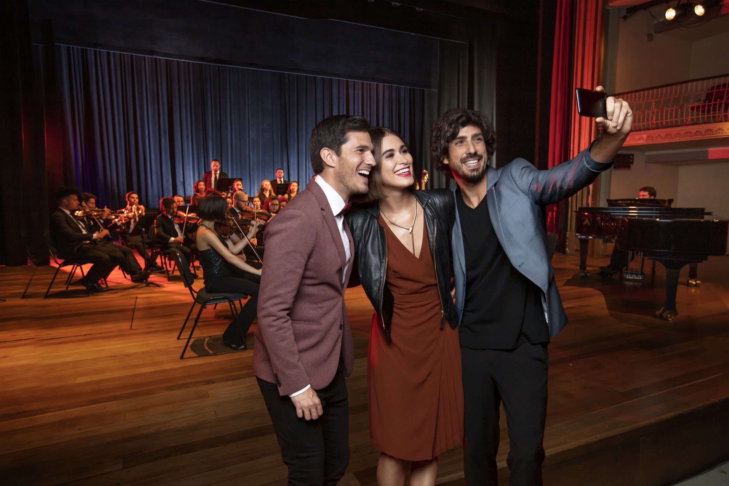Uma mulher e dois homem tiram uma selfie em um concerto musical. O homem à direita tem cabelos pretos, barba e veste um blêizer cinza, camiseta preta e calça preta. A mulher no centro usa vestido marrom, com jaqueta de couro preta e um colar dourado. O homem à esquerda veste um blêizer de cor vermelho-escuro, camisa branca e calça preta. No fundo da imagem há instrumentistas tocando violinos à esquerda e um pianista à direita.